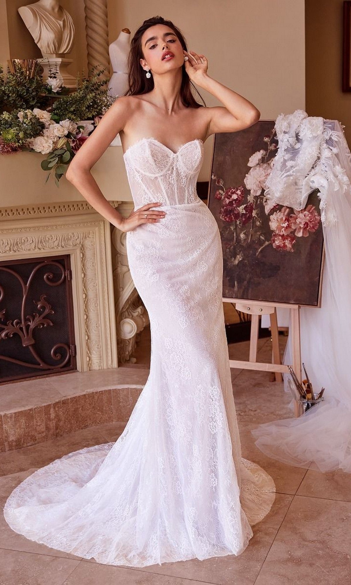 A woman in the Andrea and Leo WL040 long wedding dress, featuring a fitted lace design and sweetheart neckline, poses indoors beside a fireplace and a floral painting on an easel.
